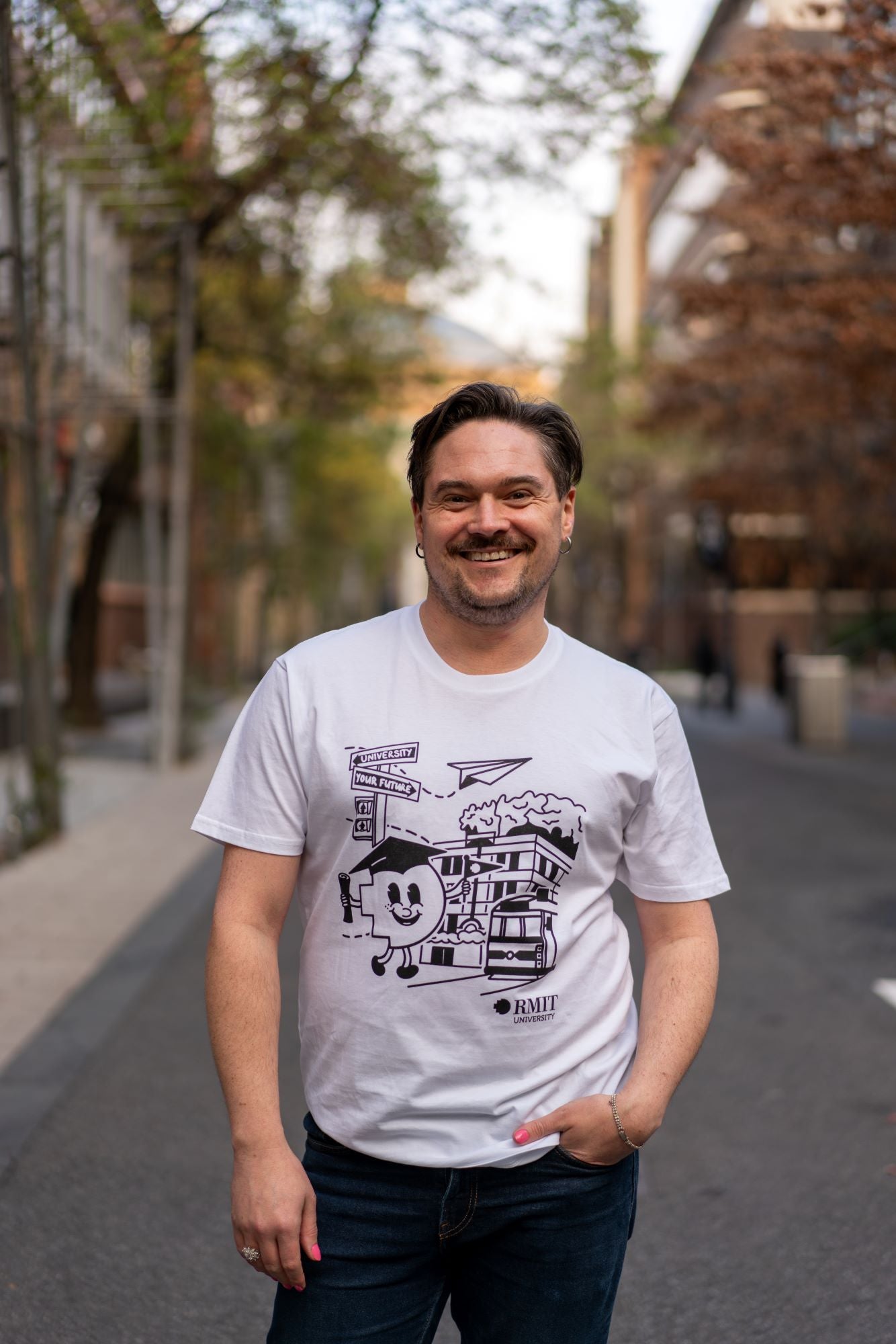Further-up front view of the RMIT Graduate Parade T-shirt worn by a smiling model facing the camera. The white tee features a black print of an animated pixel figure wearing a mortarboard in front of the RMIT building on Swanston Street, available at the RMIT Store.